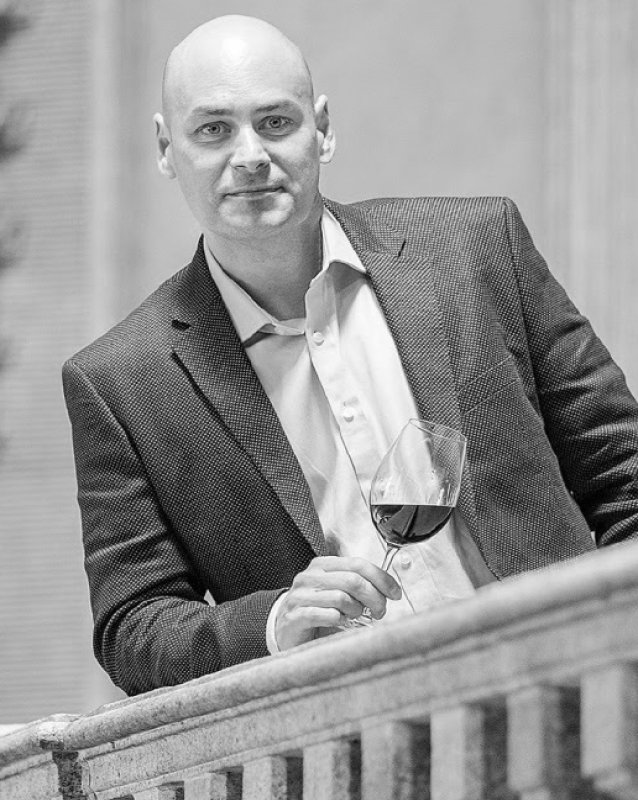 Fotografia a preto e branco de Dirceu Vianna Junior, Master of Wine, apoiado numa varanda com um copo de vinho tinto na mão