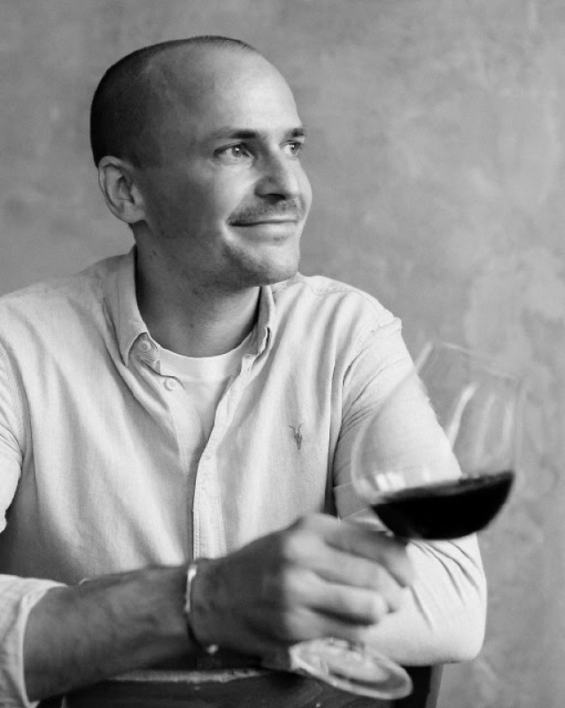 Fotografia a preto e branco de Konstantin Baum, Master of Wine, sorrindo e segurando um copo de vinho tinto